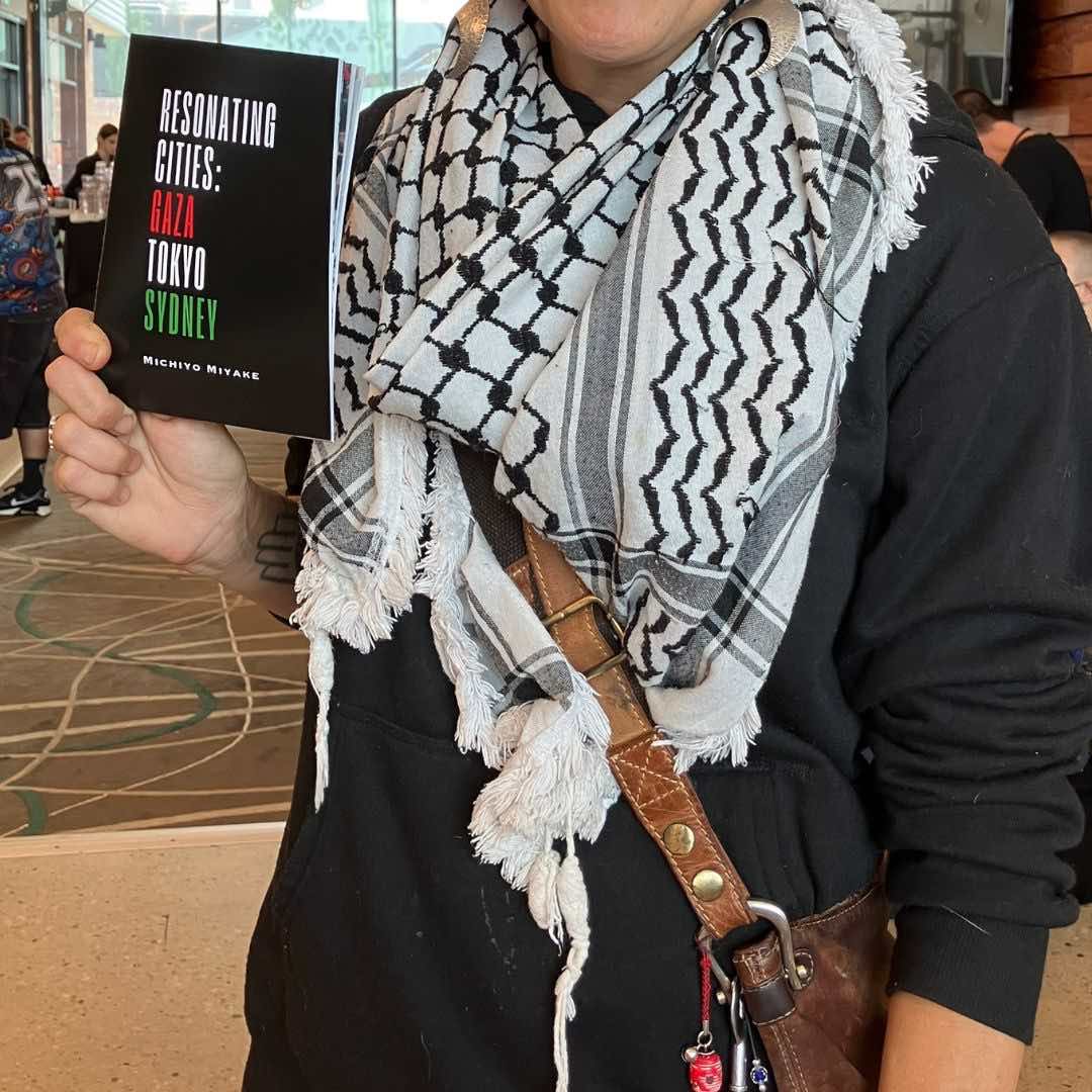 Woman wearing a Palestinian keffiyeh holding Michiyo Miyake’s poetry and visual zine Resonating Cities: Gaza, Tokyo, Sydney